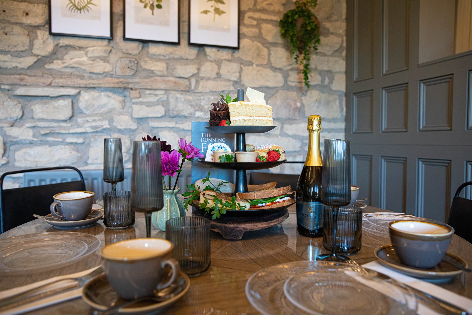 A Running Fox afternoon tea on a three-tier stand with sandwiches, scones and cakes, set with cups, glasses and a bottle of fizz against a stone wall