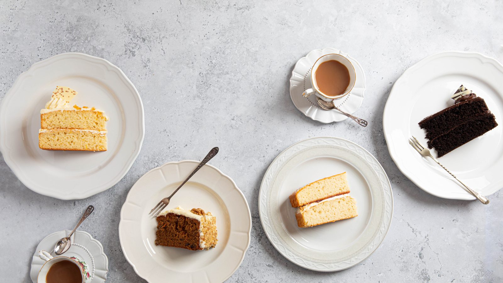 Four slices of cake, including a chocolate sponge, on white plates with cups of tea, seen from above
