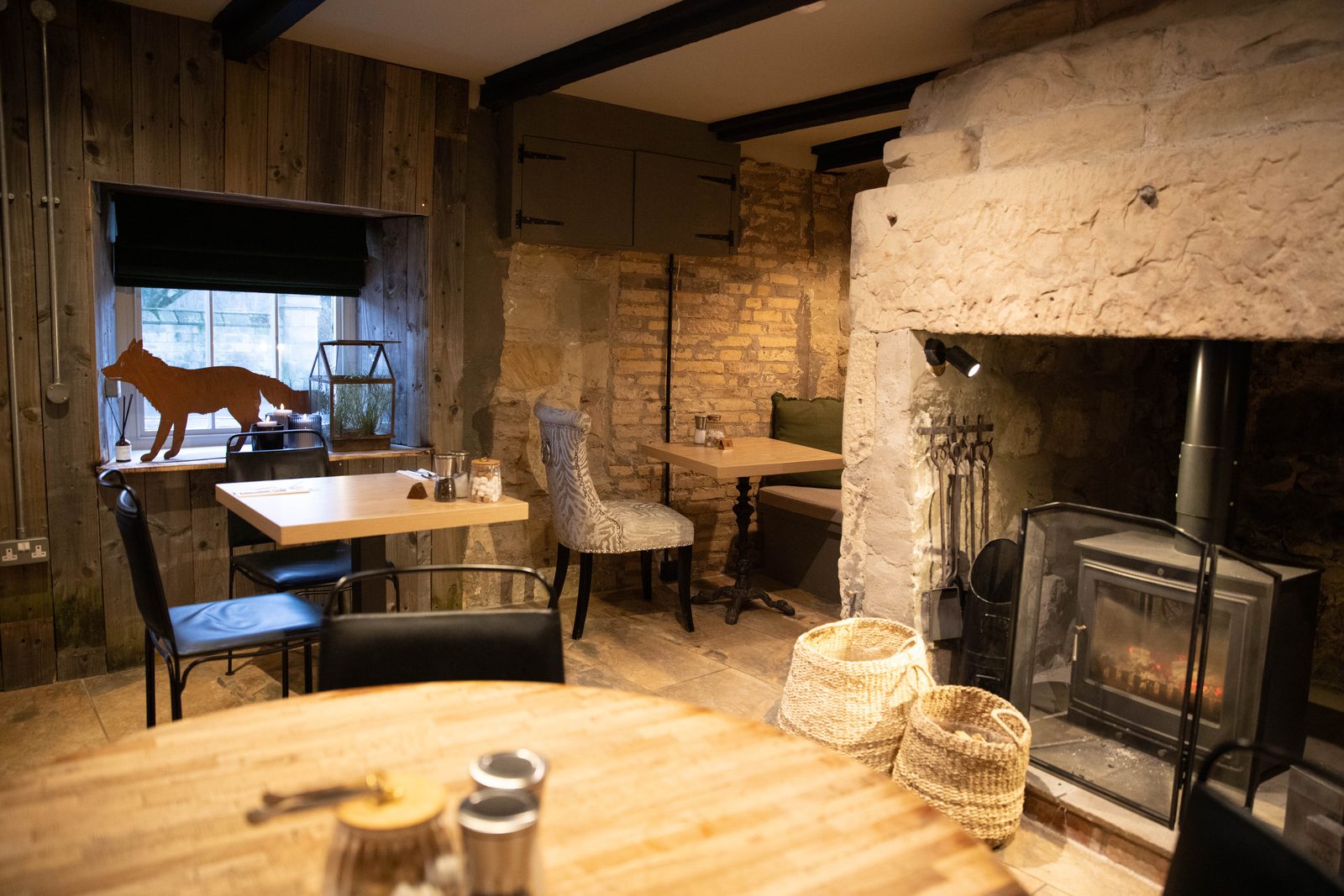 A Running Fox dining room with a wood burner set into a big stone fireplace, wicker baskets, wooden tables and a fox silhouette in the window
