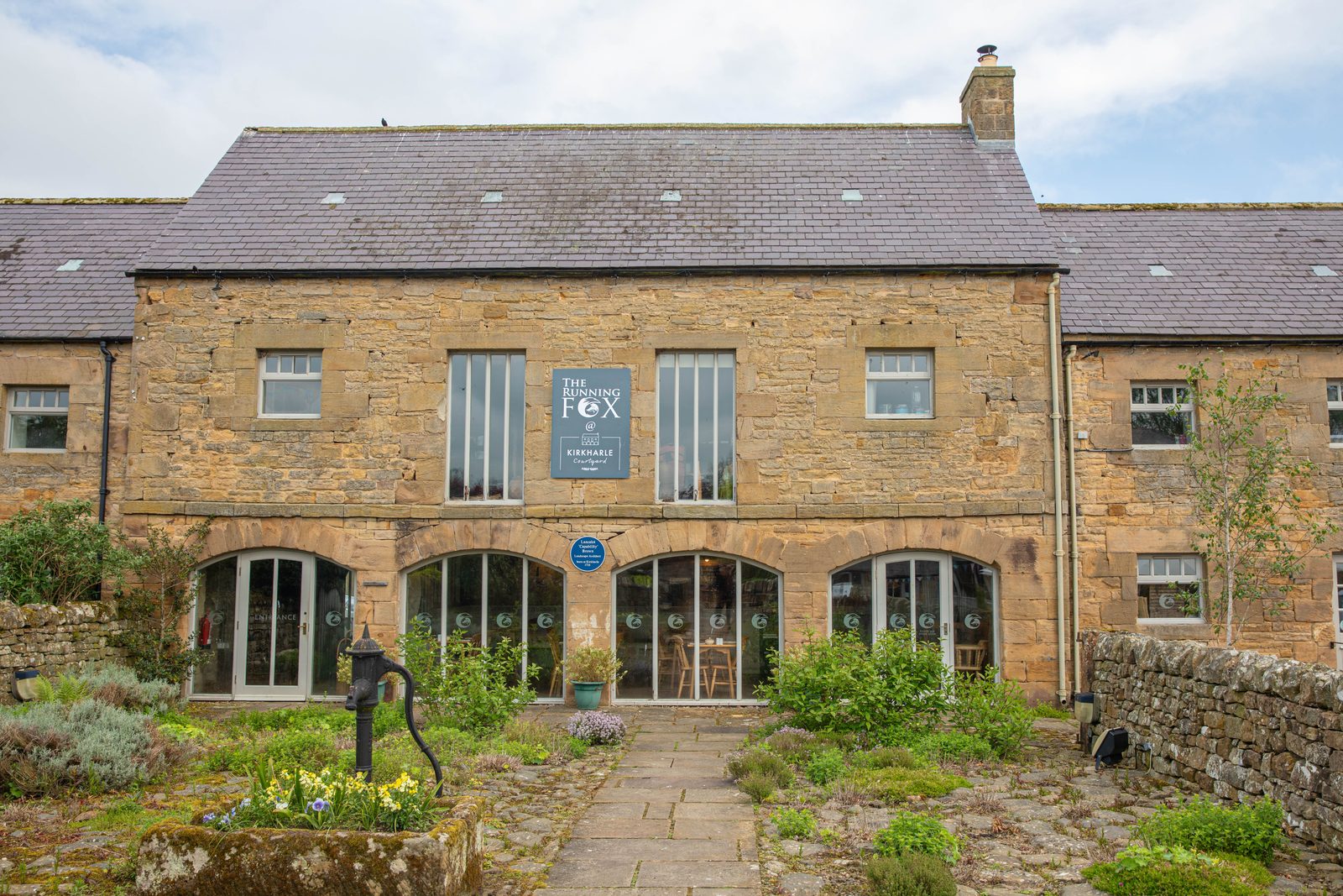 The stone front of The Running Fox at Kirkharle Courtyard, with arched windows, a hanging sign and a cottage garden path