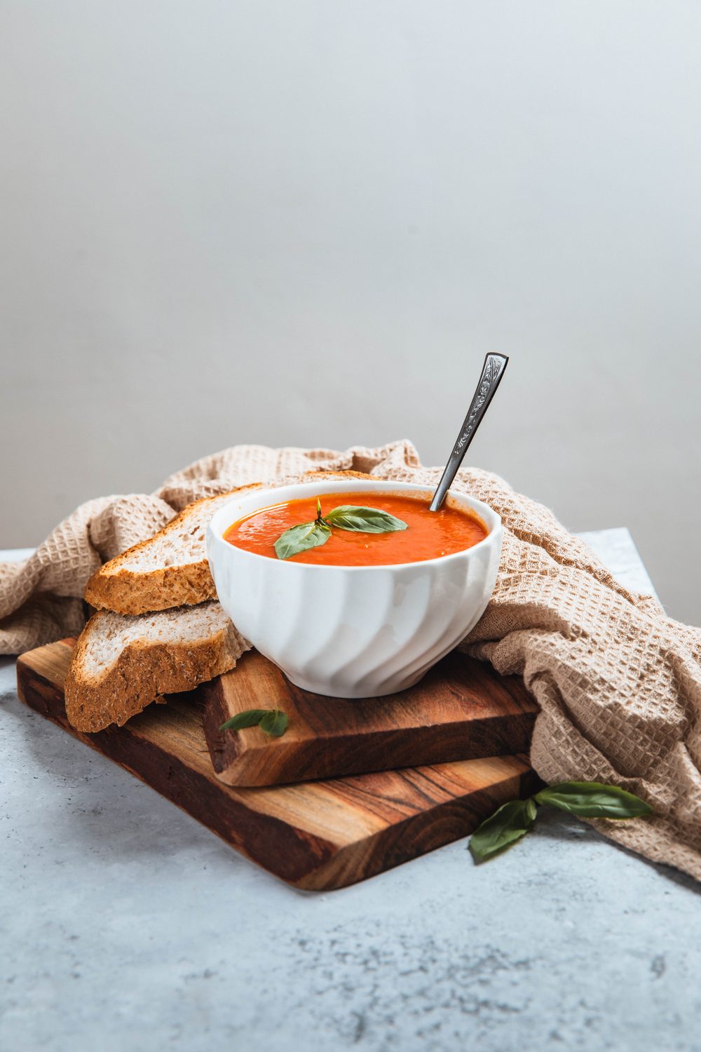 A bowl of tomato soup garnished with basil, with slices of crusty bread on a wooden board and a knitted cloth