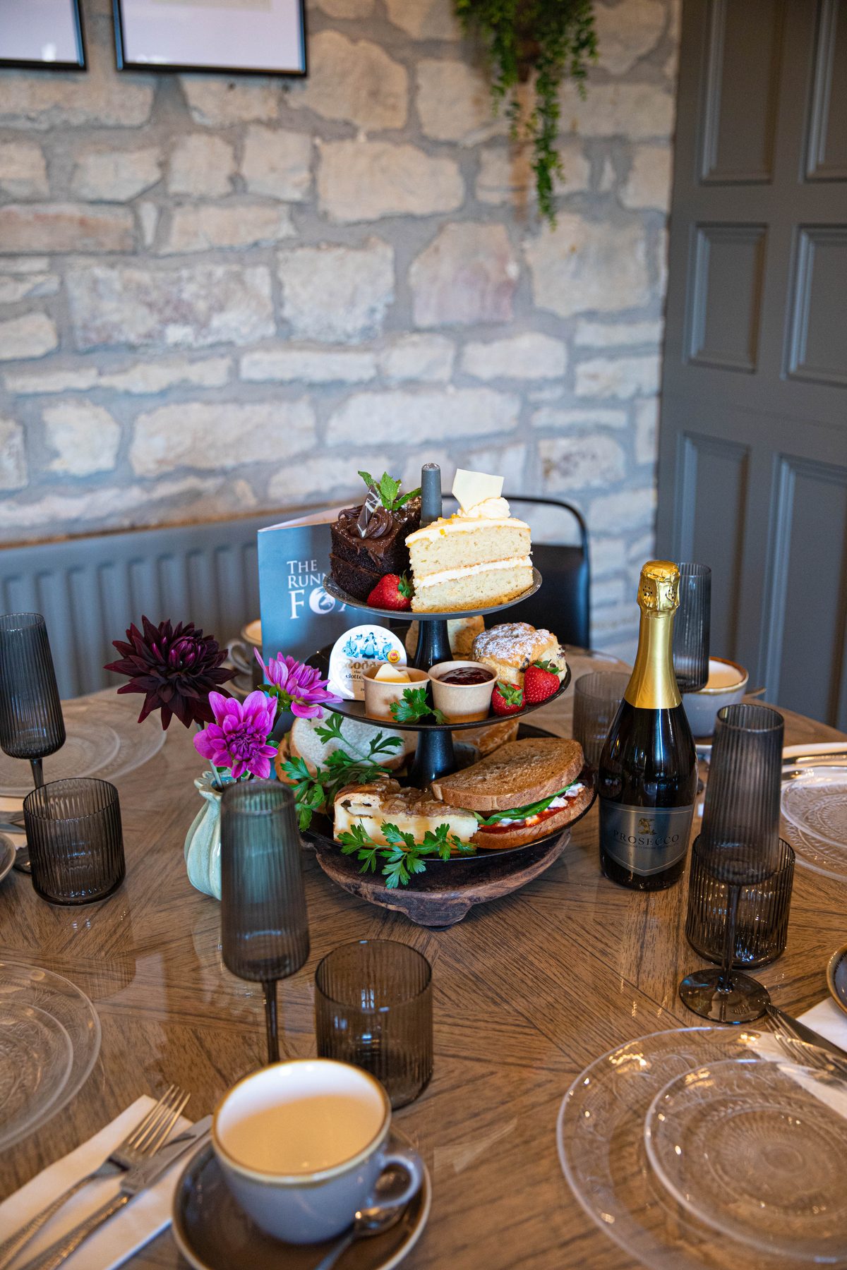 A three-tier afternoon tea stand at The Running Fox set for two, with sandwiches, scones, cake and a bottle of prosecco on a wooden table
