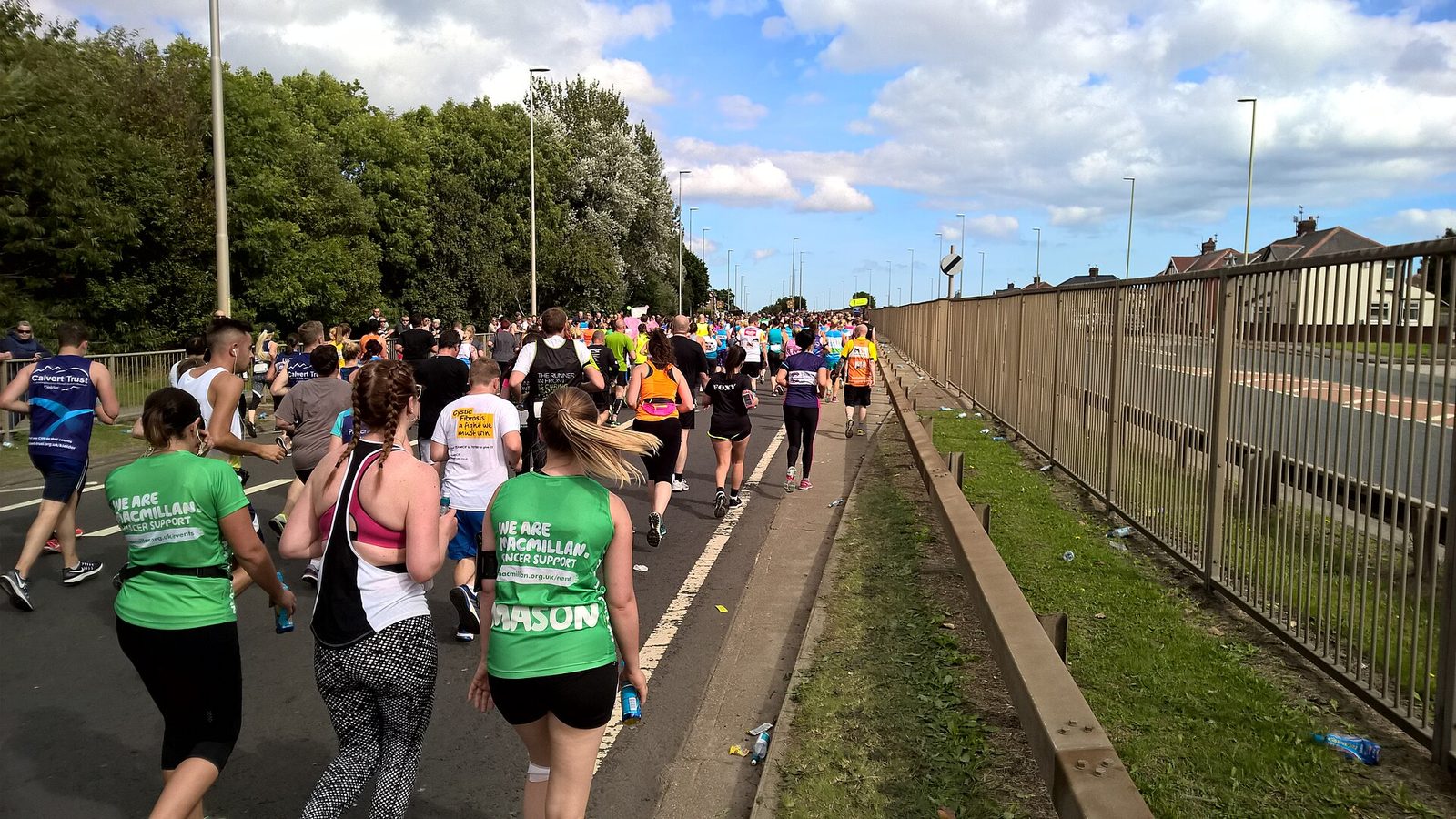 Runners seen from behind climbing a long open road on the Great North Run, several in green Macmillan charity vests, under a blue sky with white clouds