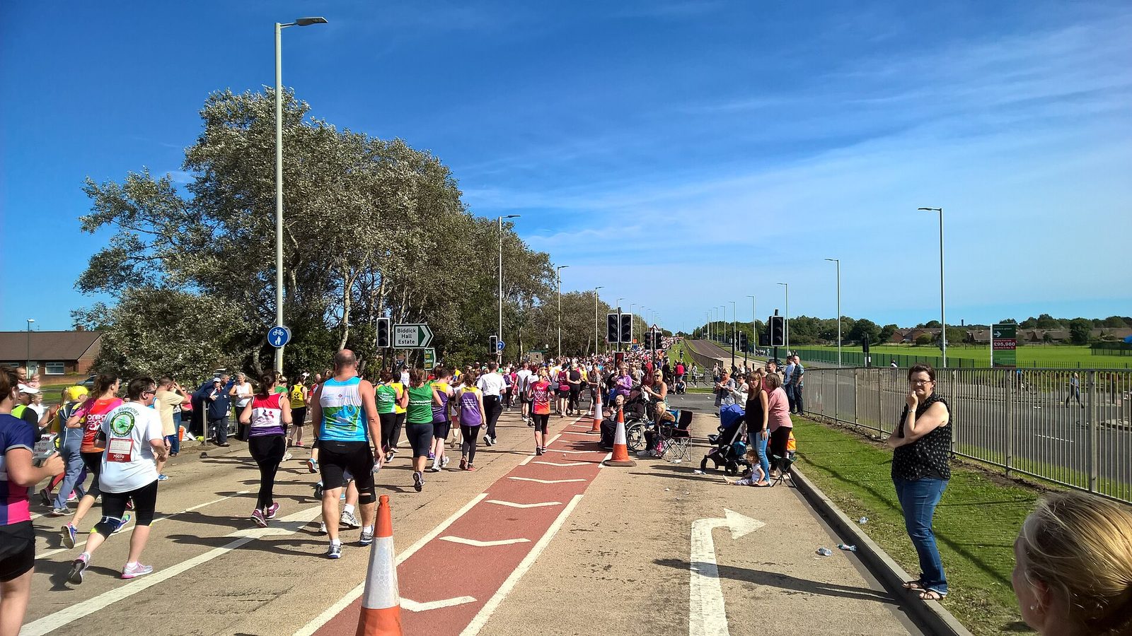 A broad sunlit road on the Great North Run route lined with spectators, with runners and walkers spread across the carriageway