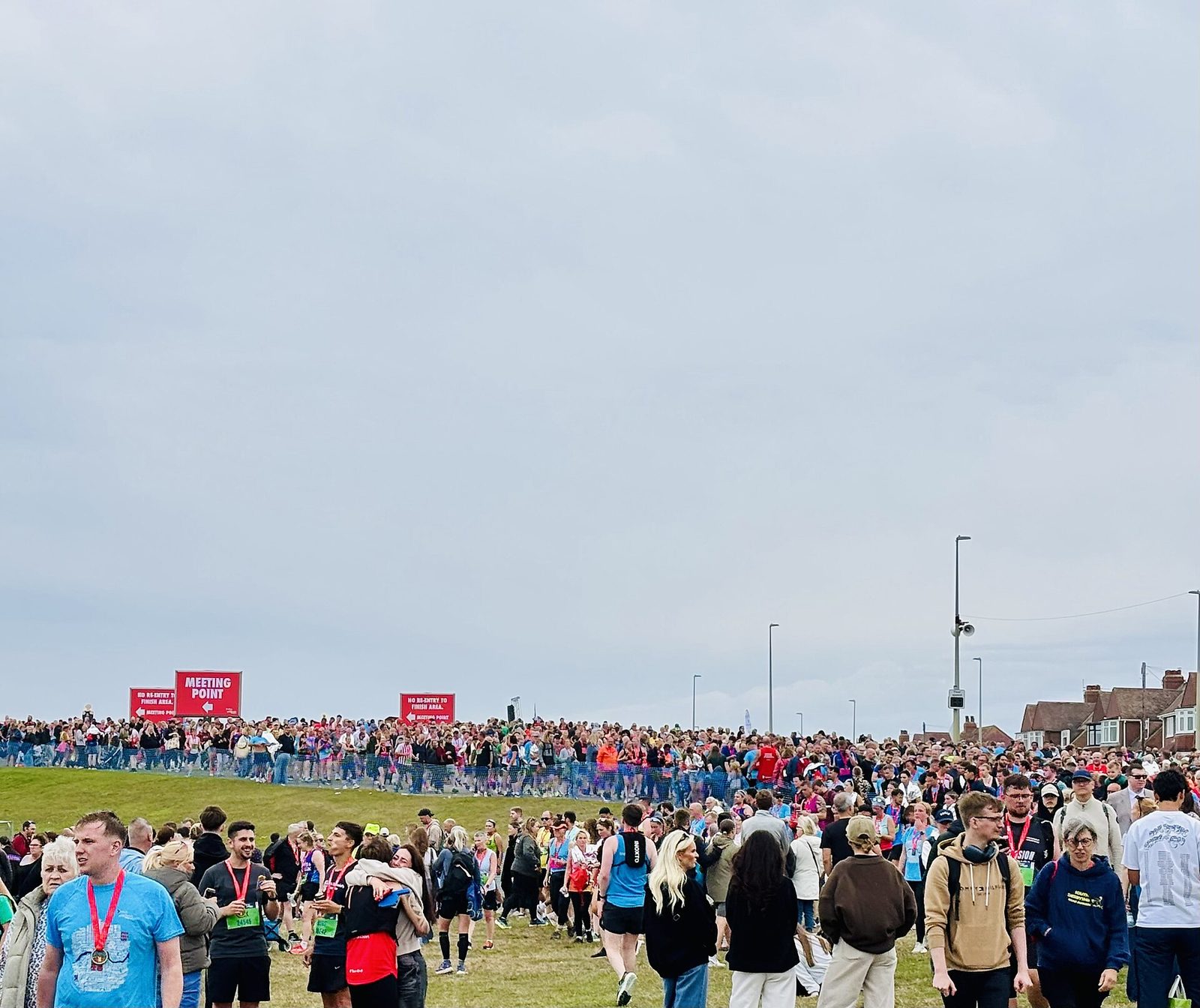 A large crowd gathered on the grass at the Great North Run finish area, many wearing finishers' medals, with meeting point signs behind them