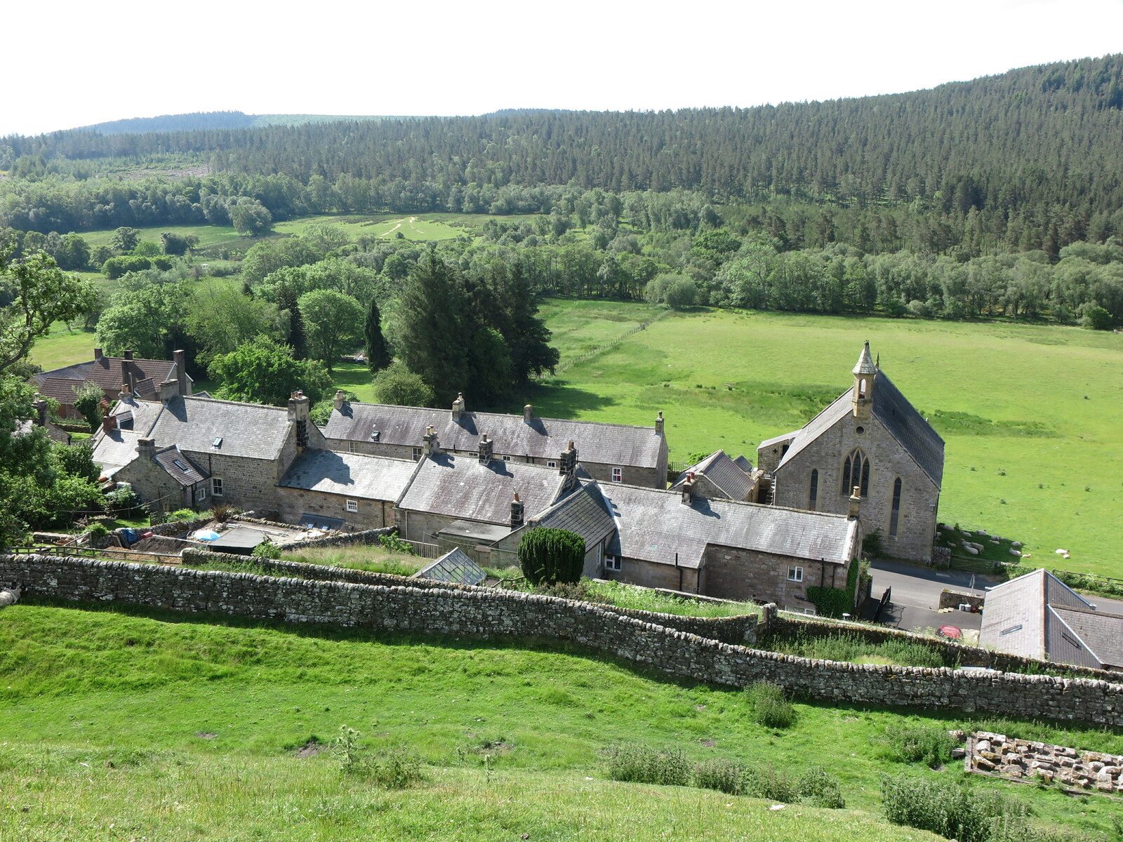 The village of Harbottle below a wooded hillside, seen from the grassy earthworks of Harbottle Castle